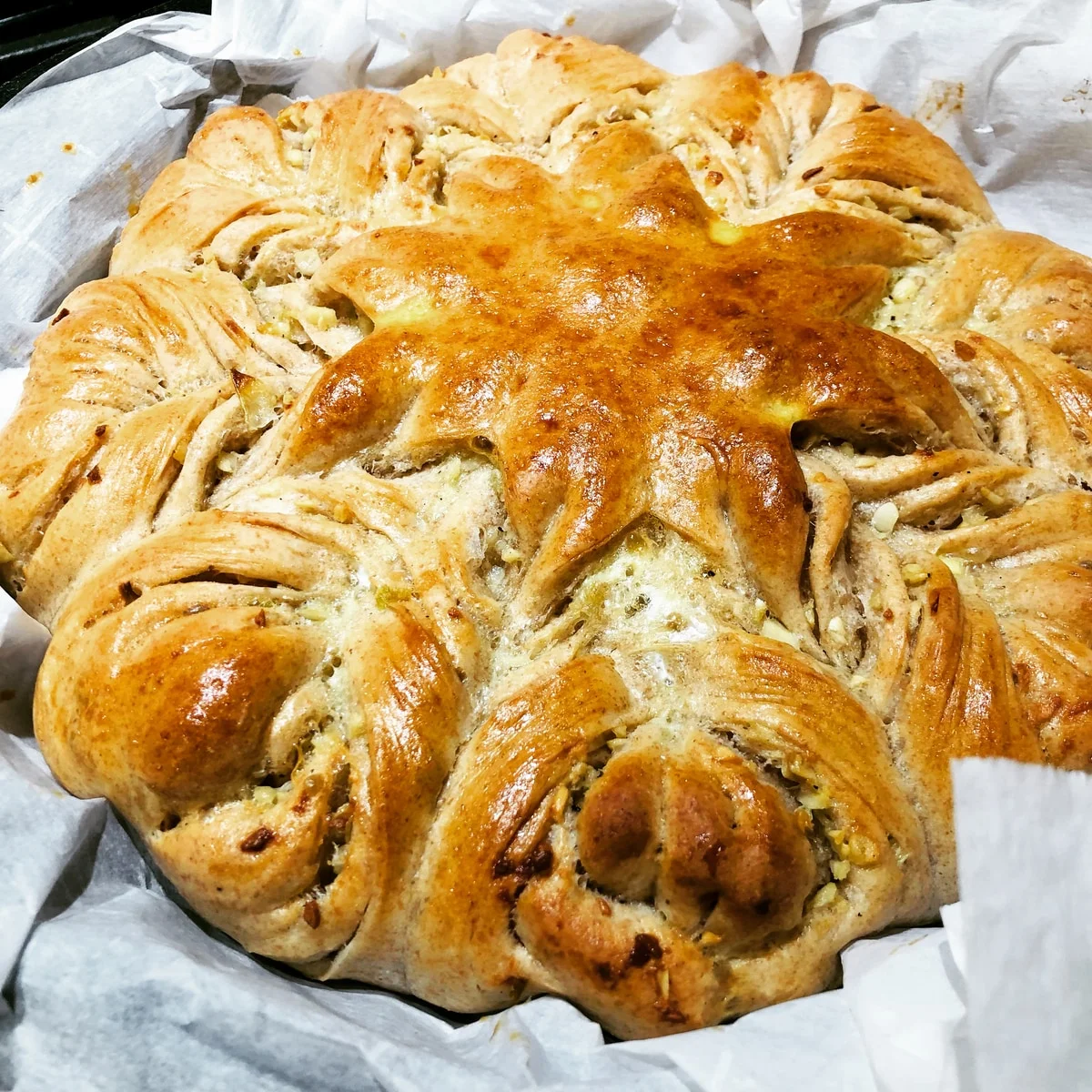 A golden round loaf with twisted sections around a flower-shaped centre, resting on baking paper.