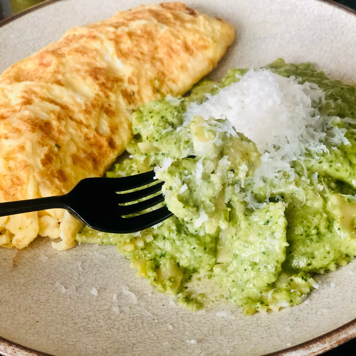 A folded golden omelette beside pasta in green sauce topped with grated cheese, with a fork lifting one piece.