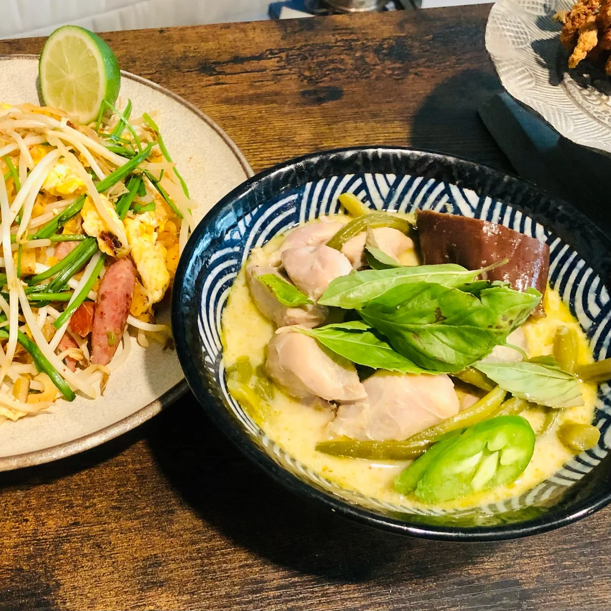 A bowl of creamy sauce with meat, vegetables, and green herbs beside noodles topped with bean sprouts and a lime slice.