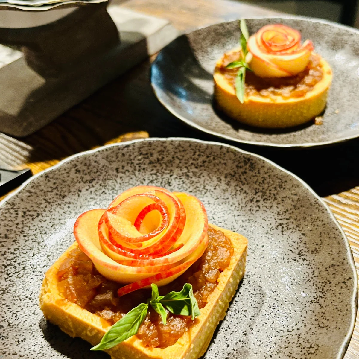 Two small tarts topped with rose-shaped fruit slices on grey plates.