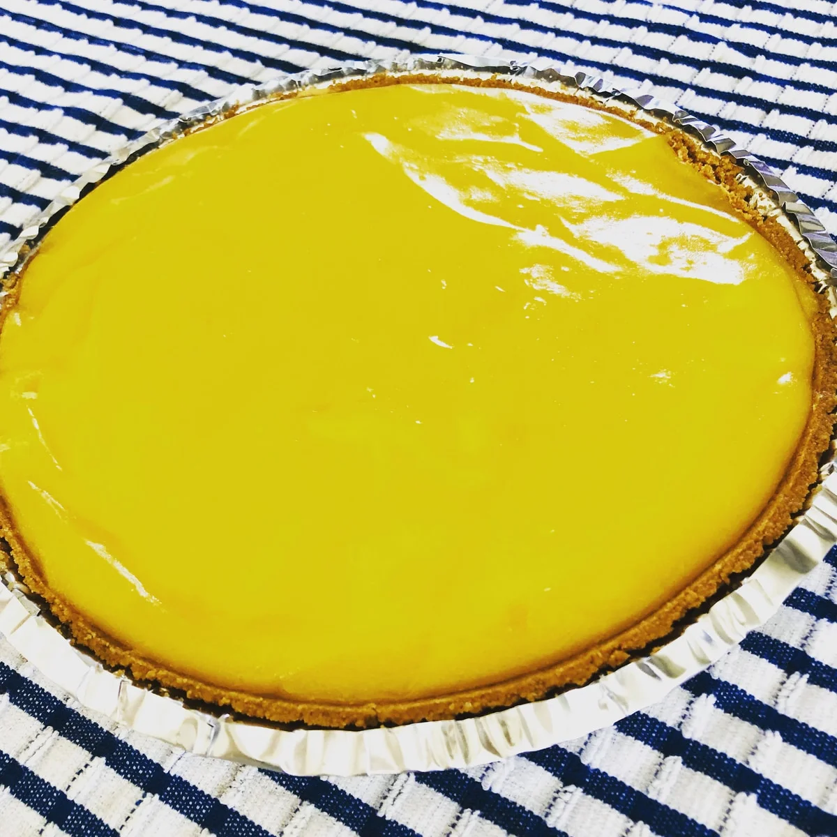A round tart with smooth yellow filling in a foil tin on a blue-and-white cloth.